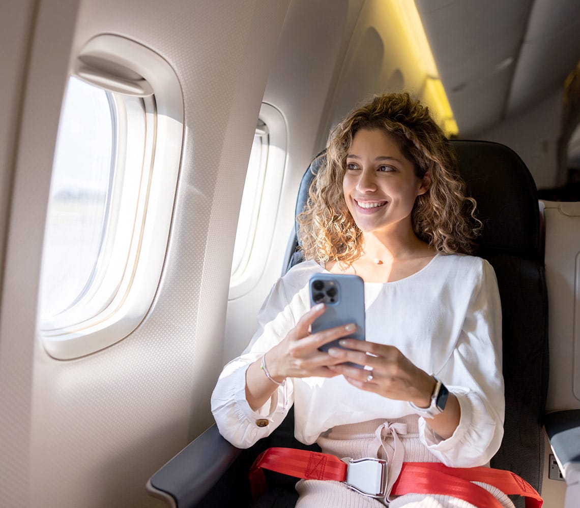 A woman seated on an airplane with phone in hand and looking out the window of the plane.